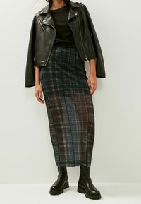 Veste en cuir noire, haut noir ajusté et longue jupe à carreaux dans des teintes de vert foncé et de marine, accompagnés de bottes noires épaisses.