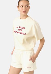 Kreminė medvilninė marškinėlių ir šortų komplektas su ryškiu raudonu tekstu: "LA MANIA 2010 PRÊT-À-PORTER" ir "LAMANIA" ant šortų. Švelni tekstūra.