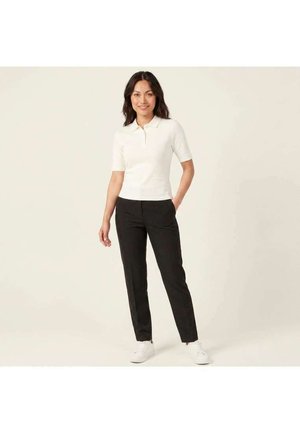Femme debout, une main dans la poche, portant un polo blanc à manches courtes, un pantalon noir et des baskets blanches sur un fond uni.