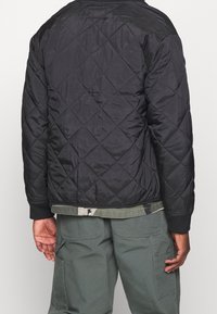 Zwarte gewatteerde bomberjack met een getextureerd oppervlak, geribbelde manchetten en een cropped pasvorm, gedragen over groene cargobroeken.