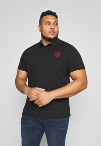 Mustapaitainen lyhythihainen polo-shirt, jossa on kaulus ja punainen brodeerattu logo rinnassa, valmistettu pehmeästä puuvillamateriaalista. Yhdistettynä sinisiin farkkuihin.