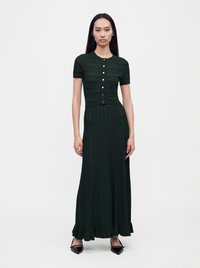 Femme aux longs cheveux noirs portant une robe en maille verte foncée à manches courtes boutonnée avec un ourlet à volants et des chaussures noires à bout pointu.