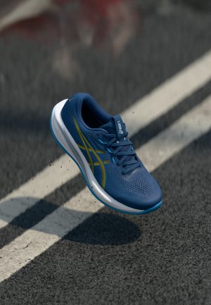 Scarpa da corsa Asics blu con strisce gialle che levita sopra una strada asfaltata con strisce bianche.