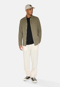 Camicia verde oliva con bottoni e tasca sul petto, t-shirt nera sotto, pantaloni chiari e sneakers scure, completati con un cappellino beige.
