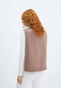 Donna con lunghi capelli rossi ricci indossa un gilet beige senza maniche sopra una maglia bianca a maniche lunghe e pantaloni bianchi, vista di spalle.