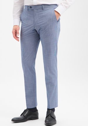 Man draagt lichtblauwe broek met patroon, zwarte nette schoenen, zwarte sokken en een wit overhemd met één hand in de zak.