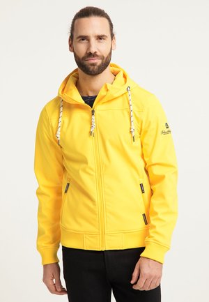 Mann mit Bart, der eine leuchtend gelbe Kapuzenjacke mit Reißverschluss und weißen Kordeln sowie schwarze Hose trägt, steht vor einem schlichten Hintergrund.
