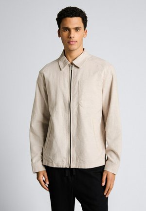 Młody mężczyzna w beżowej kurtce na zamek z kołnierzem, kieszeniami bocznymi i naszytą kieszenią na piersi, stojący na tle jasnego, jednolitego tła.