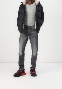 Chaqueta acolchada negra con forro interior beige, suéter gris con costillas, vaqueros grises desgastados y zapatillas negras/rojas. Enfoque en durabilidad y comodidad.