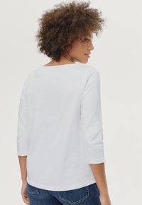 Blusa blanca de manga larga con un corte relajado, escote redondo y mangas tres cuartos, combinada con jeans azul oscuro. Textura suave de algodón.