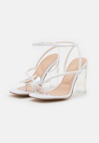 Sandali bianchi con tacchi alti trasparenti, caratterizzati da un'abbellimento scintillante e sottili cinghie su una soletta beige.