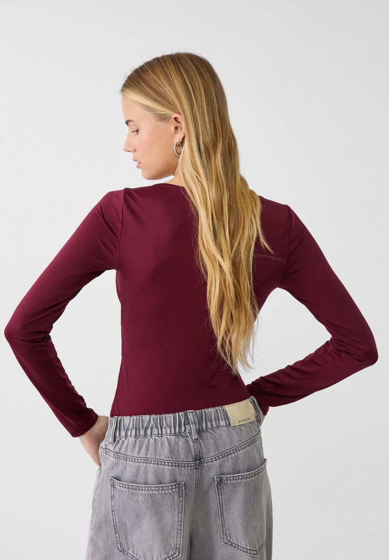 Donna con lunghi capelli biondi, che indossa orecchini a cerchio, un top fitted a maniche lunghe color bordeaux e dei pantaloni in denim grigi a vita alta, in piedi con le mani sui fianchi.