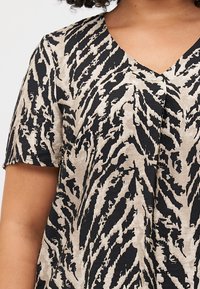 Un gros plan sur une personne portant une chemise à manches courtes avec un fond beige et un motif animal noir, dotée d'un col en V et de boutons.