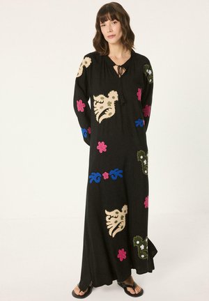 Kobieta ubrana w długą czarną sukienkę z wielokolorowymi kwiatowymi i abstrakcyjnymi wzorami, stojąca z rękami za plecami na jednolitym tle.