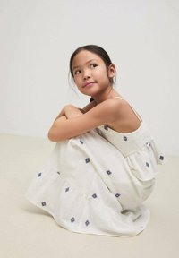 Robe en coton blanc avec broderie bleue, ourlet volanté et bretelles spaghetti. La robe a un buste smocké et tombe gracieusement.