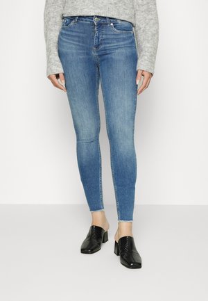 Jeans skinny azuis feitos de denim, apresentam uma cintura alta, design desbotado e bainha crua. Combinados com mules de salto texturizados pretos.