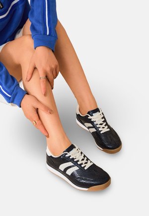 Personne portant des baskets noires et blanches avec des semelles en caoutchouc naturel, un haut de survêtement bleu, tenant et montrant son tibia gauche.