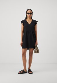 Vestido preto curto com decote em V, mangas em babados e padrão de ilhós. Combinado com sandálias pretas e uma pequena mala bege. Textura suave.