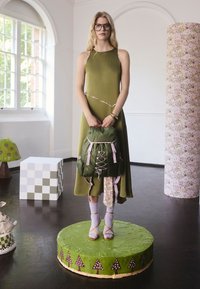 Olivovo zelené šaty s kvetinovými akcentmi, kombinované so zeleným batohom s ružovými popruhmi, ktorý je položený na zelenom kruhovom podstavci.