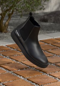 Bota chelsea de couro preto com laterais elásticas e um puxador traseiro, colocada sobre uma superfície com padrão de pedra e madeira. Apresenta uma marcação em prata.