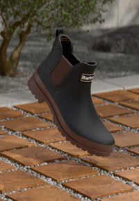 Zwarte rubberen enkelboot met een bruine elastische zijkant en logo. Textuurzool met opvallende noppen. Vloeiend boven een bakstenen oppervlak.