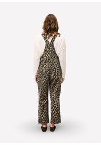 Persoon met lang krullend haar, gekleed in een crèmekleurig shirt met lange mouwen en een overall met luipaardprint, staand met zijn rug naar de camera op een witte achtergrond.