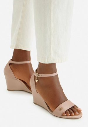 Fødder iført beige kilehælssandaler med ankelstropper og små gyldne charms, kombineret med hvide korte bukser.