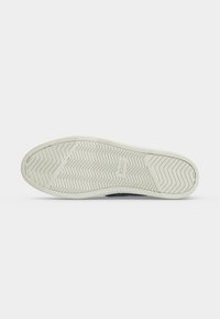 Semelle d'une chaussure en caoutchouc de couleur claire avec des motifs à chevrons et en zigzag, et le nom de la marque "TOMS" embossé au centre.