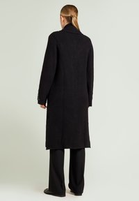 Zwarte wollen jas met een lang, gestroomlijnd ontwerp, voorzien van een grote kraag en versierde manchetten. Gecombineerd met zwarte broek met wijde pijpen.