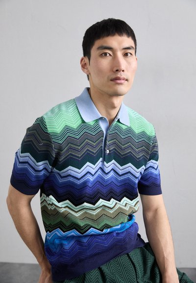 Polo cu mânecă scurtă, cu un model în zigzag în nuanțe de verde, albastru și bleumarin. Fabricat dintr-un amestec de bumbac, cu o guler de culoare albastru deschis și placket cu trei butoane.