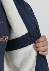 Mørkeblå zip-up jakke med blød hvid fleeceforing og en lille lynlåslomme, der har en glat ydre tekstur og brune hardwaredetaljer.