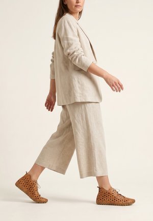 Frau in beige Leinenanzug, trägt braune Schnürschuhe mit dreieckigen Ausschnitten.