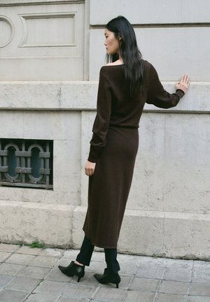 Kobieta z długimi czarnymi włosami, ubrana w brązową sukienkę midi z odkrytymi ramionami, czarne spodnie i czarne buty na obcasie, stojąca przy kamiennym murze.