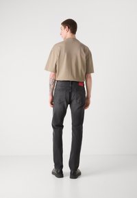 Beige short-sleeve shirt and black jeans with a red logo patch at the back. Slim fit, smooth texture. Black footwear completes the look.