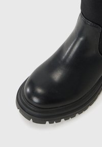 Gros plan sur une botte noire brillante avec un bout rond et une semelle épaisse et texturée, placée sur un fond gris clair.