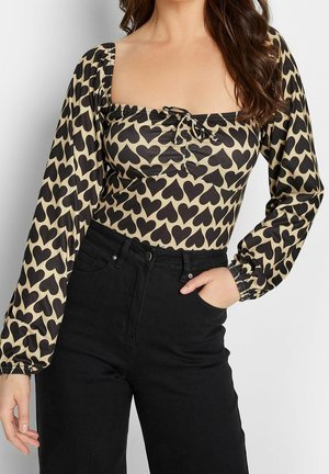 Kobieta w beżowej bluzce z długim rękawem z czarnym wzorem serc oraz wysokim stanem czarnych dżinsów, jedna ręka w kieszeni, stoi na białym tle.