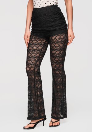 Kobieta nosząca czarne koronkowe dzwony z jednolitym panelem z wysokim stanem, w połączeniu z czarnymi sandałami na obcasie i białą bluzką w kropki.