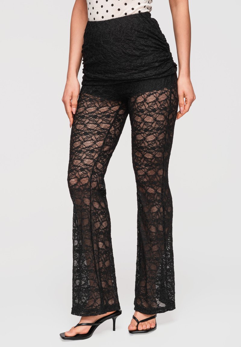 Femme portant un pantalon évasé en dentelle noire avec un empiècement taille haute uni, associé à des sandales noires à talons et un haut blanc à pois.