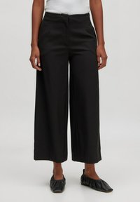 Schwarze, weit geschnittene Hosen mit glatter Textur, strukturiertem Bund und klassischen Taschen, kombiniert mit schwarzen flachen Schuhen.