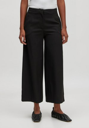Pantalons noirs à jambes larges avec une texture lisse, une taille structurée et des poches classiques, associés à des chaussures plates noires.