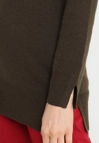 Manica di un maglione marrone a maglia con polsino a coste, caratterizzato da una piccola fessura, indossato con pantaloni rossi.