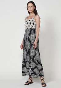 Robe maxi avec un motif damassé noir et blanc, un corsage en crochet, des bretelles spaghetti et une jupe fluide. Portée avec des sandales noires à lanières.