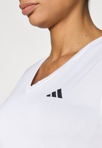 Camiseta blanca de cuello en V hecha de tela ligera con un logo negro que presenta tres franjas diagonales en el lado izquierdo.