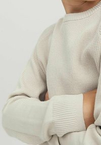 Person klädd i en krämfärgad ribbad tröja med armarna i kors, som visar det strukturerade stickade tyget och de rullade ärmsluten.