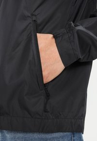 Svart lättviktig jacka med dragkedjeförsedda sidofickor. Har en slät yta och elastiska ärmslut. Avslappnad design med dragsko i nederkant.