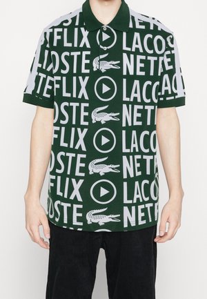 Ciemnozielona koszulka polo z białym, wyraźnym nadrukiem na całej powierzchni z napisem "NETFLIX" i "LACOSTE" obok ikon krokodyla i przycisku odtwarzania, noszona przez osobę.
