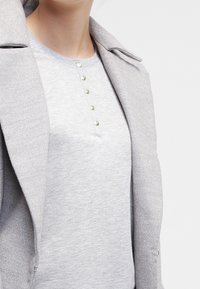 Hellgrauer Pullover mit einem gerippten Kragen und einer Knopfleiste mit goldfarbigen Knöpfen, unter einem ähnlichen grauen taillierten Mantel getragen.