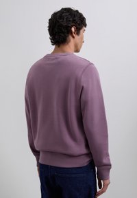 Hombre de espaldas vistiendo un sudadera de manga larga color malva y jeans oscuros frente a un fondo gris liso.