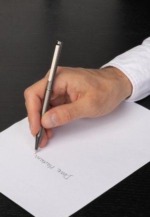 Rechterhand houdt een zilveren pen vast en schrijft "Geachte mevrouw" op wit papier boven een zwart houten bureau.