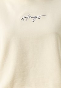 T-shirt beige clair avec une texture côtelée, arborant un logo brodé en argenté de la signature "Hugs" sur le côté supérieur gauche.